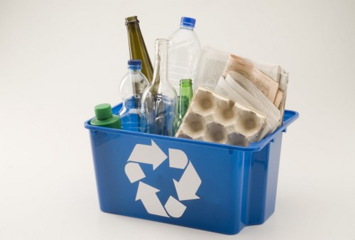 A blue plastic recycling bin filled with various types of empty bottles including glass and plastic, as well as a carton of egg boxes and crumpled newspapers, sitting on a plain light-colored surface against a neutral background. The bottles include clear, green, and brown glass with some caps still attached. The packaging materials and newspapers indicate waste suitable for rubbish removal services in Heston, with the bin featuring a white recycling symbol on the side. The arrangement of the waste appears stacked and slightly overflowing, suitable for collection or clearance, typical of garden or household rubbish clearance tasks performed by Garden Clearance Heston. The scene emphasizes disposal of recyclable items and paper waste, aligned with local rubbish collection or clearance services.
