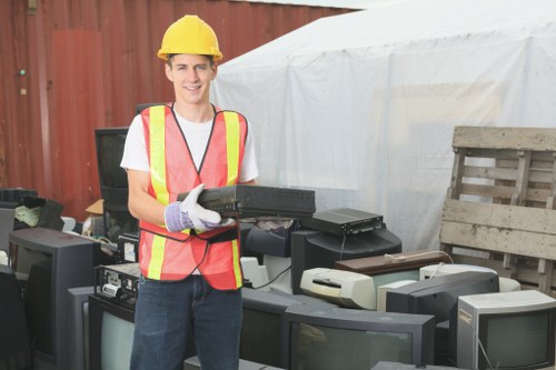 A young male worker wearing a yellow safety helmet, a high-visibility pink and yellow vest, and protective gloves is standing outdoors next to a collection of discarded electronic devices, including old computer monitors and a printer, placed on a work surface. The background features a wooden fence and a white tent or canopy structure, indicating an outdoor storage or disposal area for electronic waste. The worker is smiling and appears to be preparing to load or move the electronic rubbish, which is in line with rubbish removal services that dispose of unwanted or obsolete electrical items. The setting suggests a professional environment related to waste collection or clearance, possibly in or near Heston, with a focus on responsible electronic waste disposal. Garden Clearance Heston provides such services, ensuring safe and proper removal of electronic rubbish from residential or commercial sites.