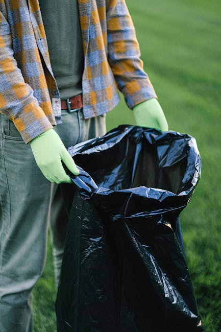 A person wearing a plaid shirt in shades of yellow, grey, and blue, blue jeans, and bright green gloves is holding open a large black rubbish bag made of shiny plastic material. The bag is partially f