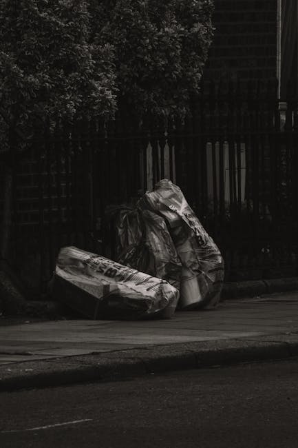 A person wearing a plaid shirt in shades of yellow, grey, and blue, blue jeans, and bright green gloves is holding open a large black rubbish bag made of shiny plastic material. The bag is partially f