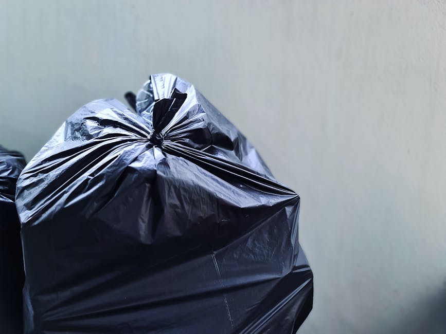 Side view of a small, aged blue pickup truck parked along the street, loaded with a large amount of mixed waste materials including flattened cardboard boxes, black plastic bags, and various household