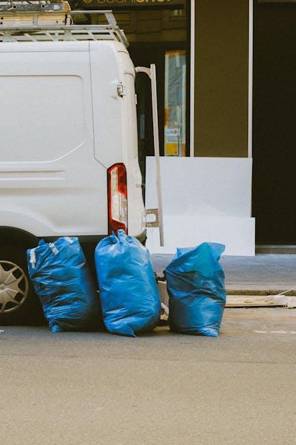 A pile of discarded cardboard boxes, some flattened and others partially assembled, along with plastic packaging and a large textile sack, all situated on uneven ground near a rough brick and concrete