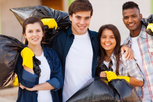 A group of four young adults standing outdoors on a paved residential street in Heston, with two women and two men smiling at the camera, each wearing yellow work gloves and holding large black rubbish bags filled with waste, indicating participation in a community cleanup or rubbish removal activity. The background features a row of houses with front gardens, some greenery, and a partly cloudy sky, suggesting a typical suburban environment. The image captures a sense of teamwork and environmental responsibility related to rubbish clearance services, such as those offered by Garden Clearance Heston, with visual emphasis on the collection of waste materials in preparation for disposal or recycling. The scene is brightly lit with natural daylight, highlighting their cheerful expressions and the contents of the rubbish bags, which appear to contain garden debris or household waste. This image reflects community engagement in rubbish removal and garden clearance activities in the local area, aligning with professional services for domestic and garden waste collection in Heston and surrounding areas within postcode vicinity.
