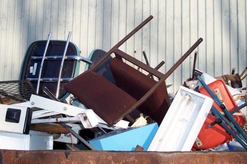 A large pile of discarded household and garden items stacked outdoors against a corrugated metal wall, including a brown upholstered chair with wooden legs, a white plastic window frame, a red plastic chair, and various metal or plastic garden tools. The items are arranged haphazardly, with some partially overlapping and leaning on each other, on what appears to be a backyard or driveway area. The collection of rubbish is typical of a garden clearance or home decluttering service that might be offered by Garden Clearance Heston, indicative of waste removal solutions in the Heston area postcode. The scene suggests an ongoing or completed rubbish removal activity, emphasizing clutter clearing that professional services can address for residents or businesses in west London. The lighting appears natural, casting soft shadows, and the environment looks clean but cluttered, ready for collection or disposal as part of garden clearance or general rubbish removal.